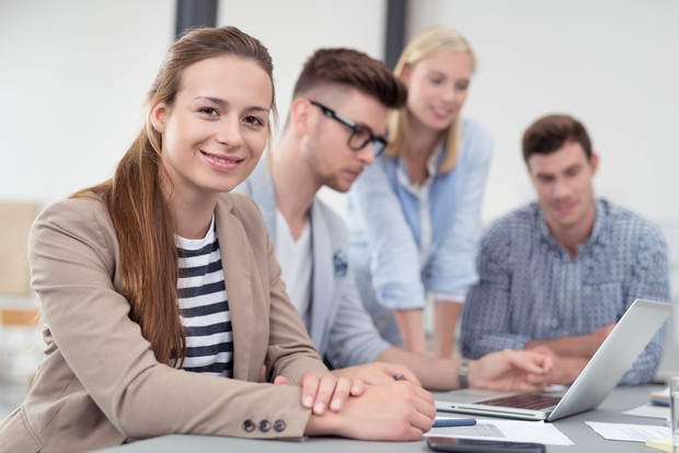 Foto: Junge Frau lächelt in die Kamera, im Hintergrund drei ArbeitskollegInnen (Bild: AdobeStock/Contrastwerkstatt). Foto: Junge Frau lächelt in die Kamera, im Hintergrund drei ArbeitskollegInnen (Bild: AdobeStock/Contrastwerkstatt).